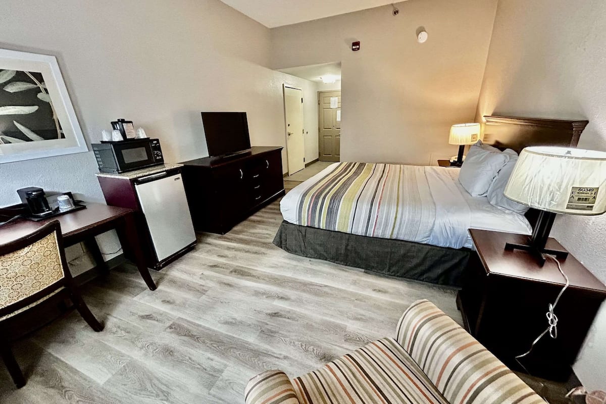 A hotel room with a bed, a desk with a computer and a coffee maker, a TV, a lamp, and a striped sofa.