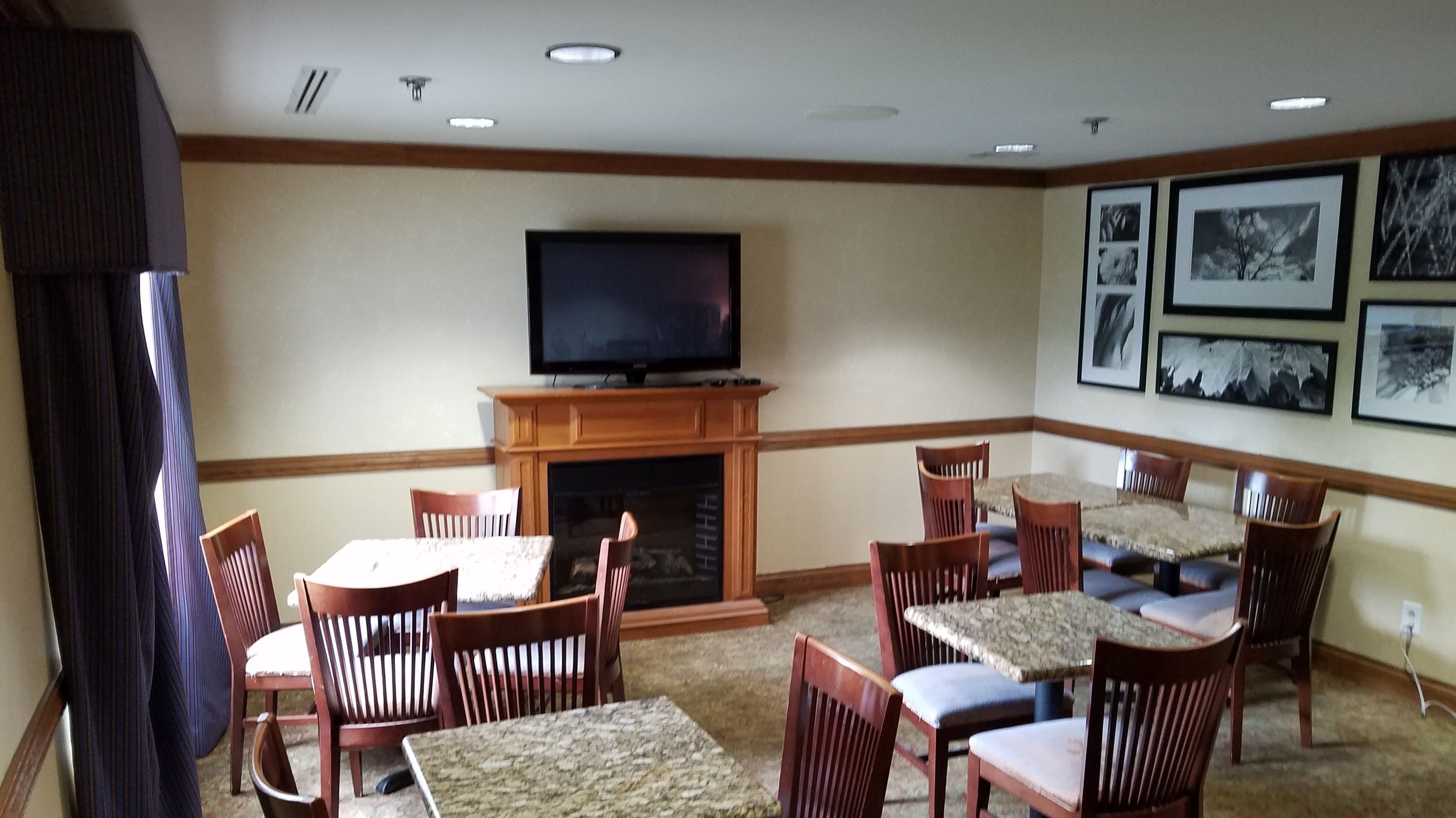 Un salón de hotel con una televisión de pantalla plana sobre una chimenea, rodeado de sillas y mesas de madera.