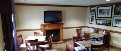 Un salón de hotel con una televisión de pantalla plana sobre una chimenea, rodeado de sillas y mesas de madera.