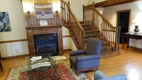 A living room with a fireplace, a blue armchair, a glass coffee table with brochures, a wooden staircase, and a framed picture on the wall.