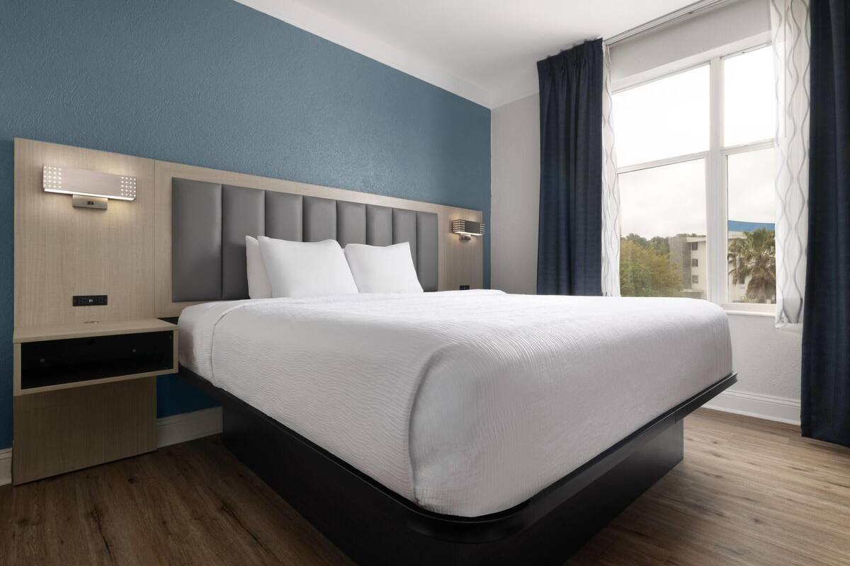 Une chambre d’hôtel moderne dotée d’un grand lit, de tables de chevet intégrées et offrant une vue sur la ville grâce à une grande fenêtre.