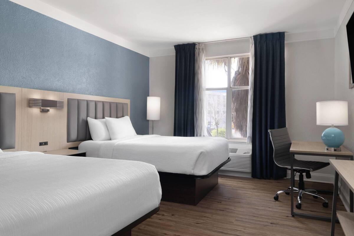 Une chambre d’hôtel avec deux lits, un bureau avec une chaise, une lampe et une fenêtre avec des rideaux.