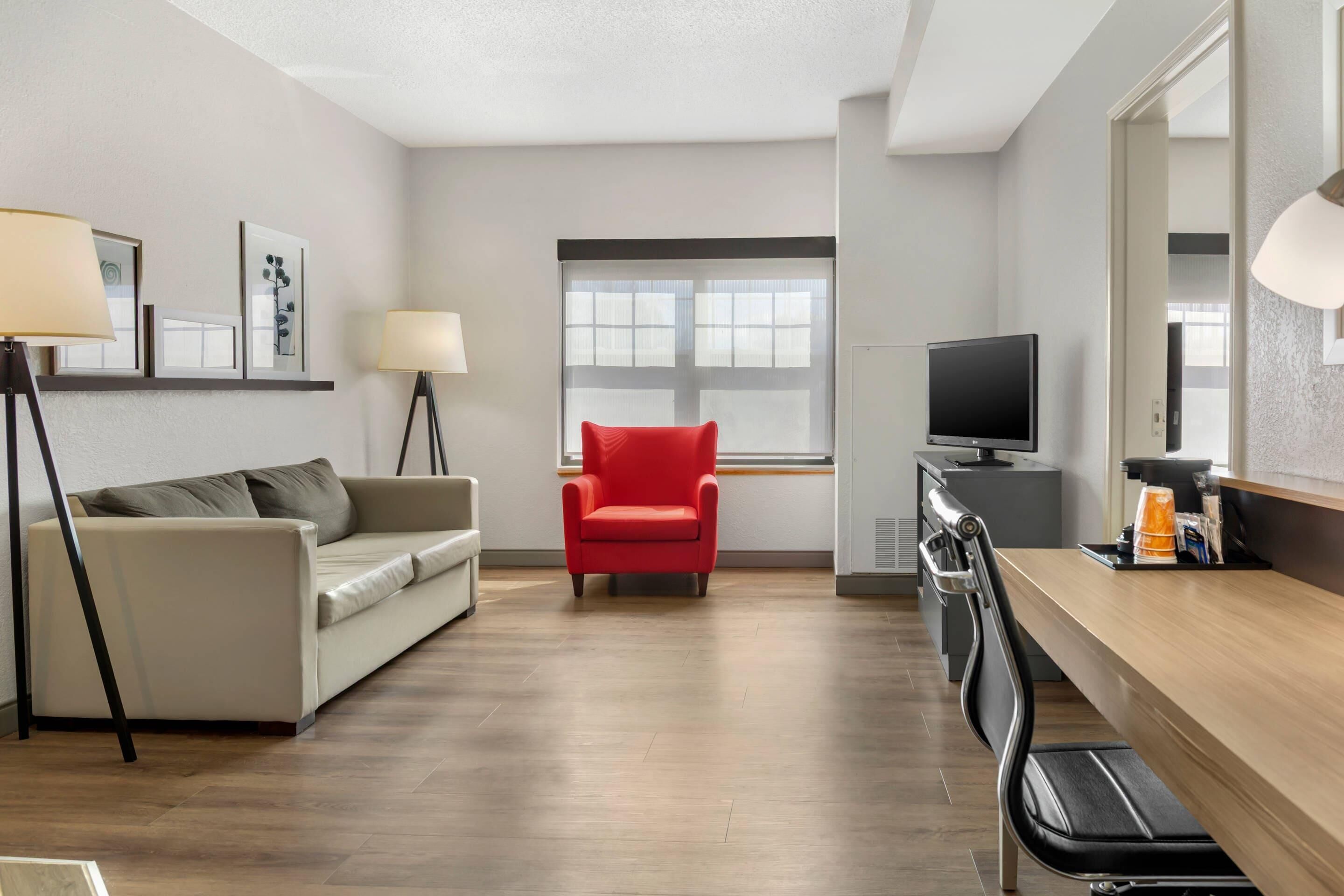 A modern hotel room with a sofa, a red chair, a TV, and a desk with a lamp.