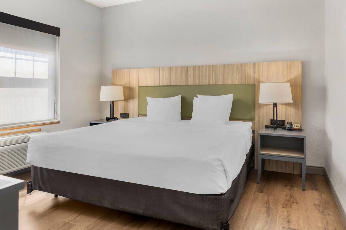 A hotel room with a large bed, two bedside tables with lamps, a wooden headboard, and a window with blinds.