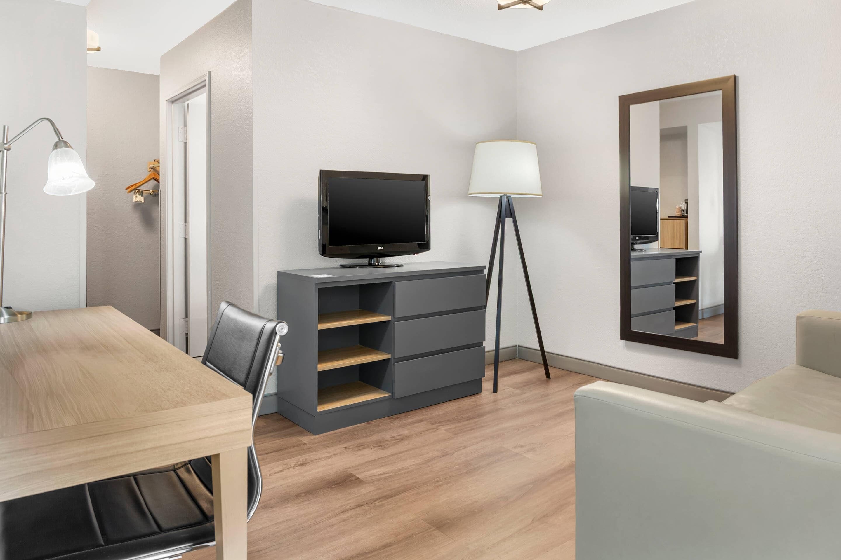 Habitación de hotel moderna con televisión de pantalla plana, un escritorio con silla, una lámpara, un espejo y estanterías empotradas.