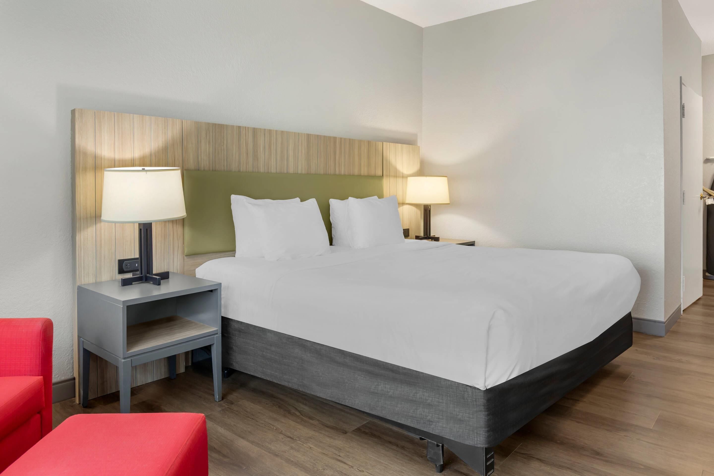 A hotel room with a bed, two bedside tables with lamps, a red chair, and a wooden floor.