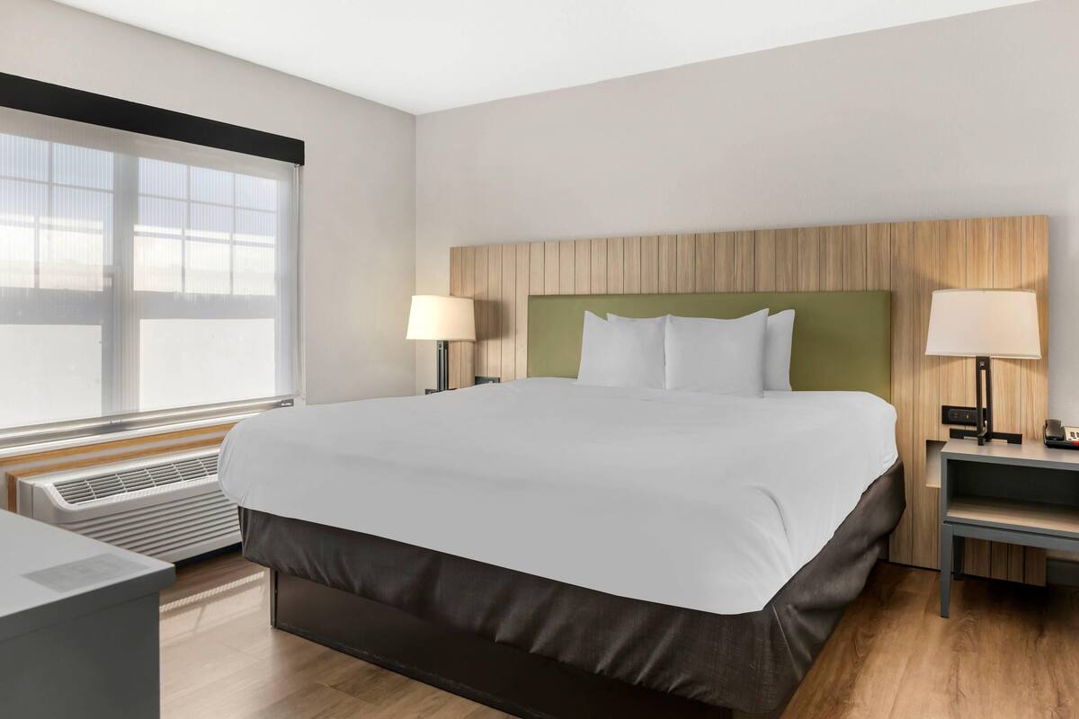 A hotel room with a large bed, two bedside tables with lamps, a window with blinds, and a wall-mounted heater.