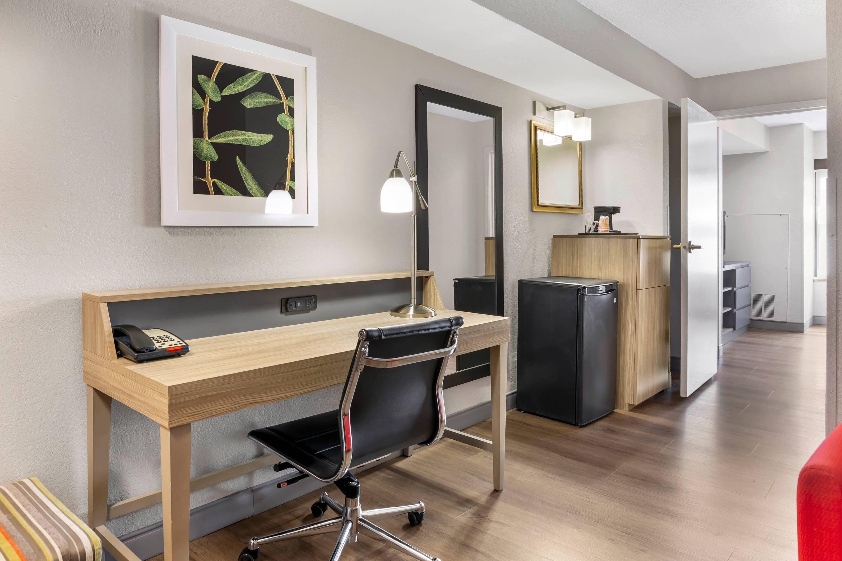 A hotel room with a wooden desk, a black office chair, a desk lamp, a refrigerator, a mirror, and a framed picture on the wall.