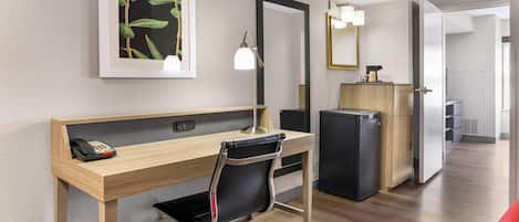 A hotel room with a wooden desk, a black office chair, a desk lamp, a refrigerator, a mirror, and a framed picture on the wall.