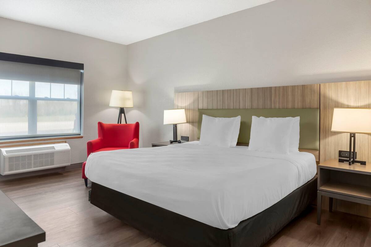 A hotel room with a large bed, two bedside tables with lamps, a red chair, a window with blinds, and a wall-mounted heater.