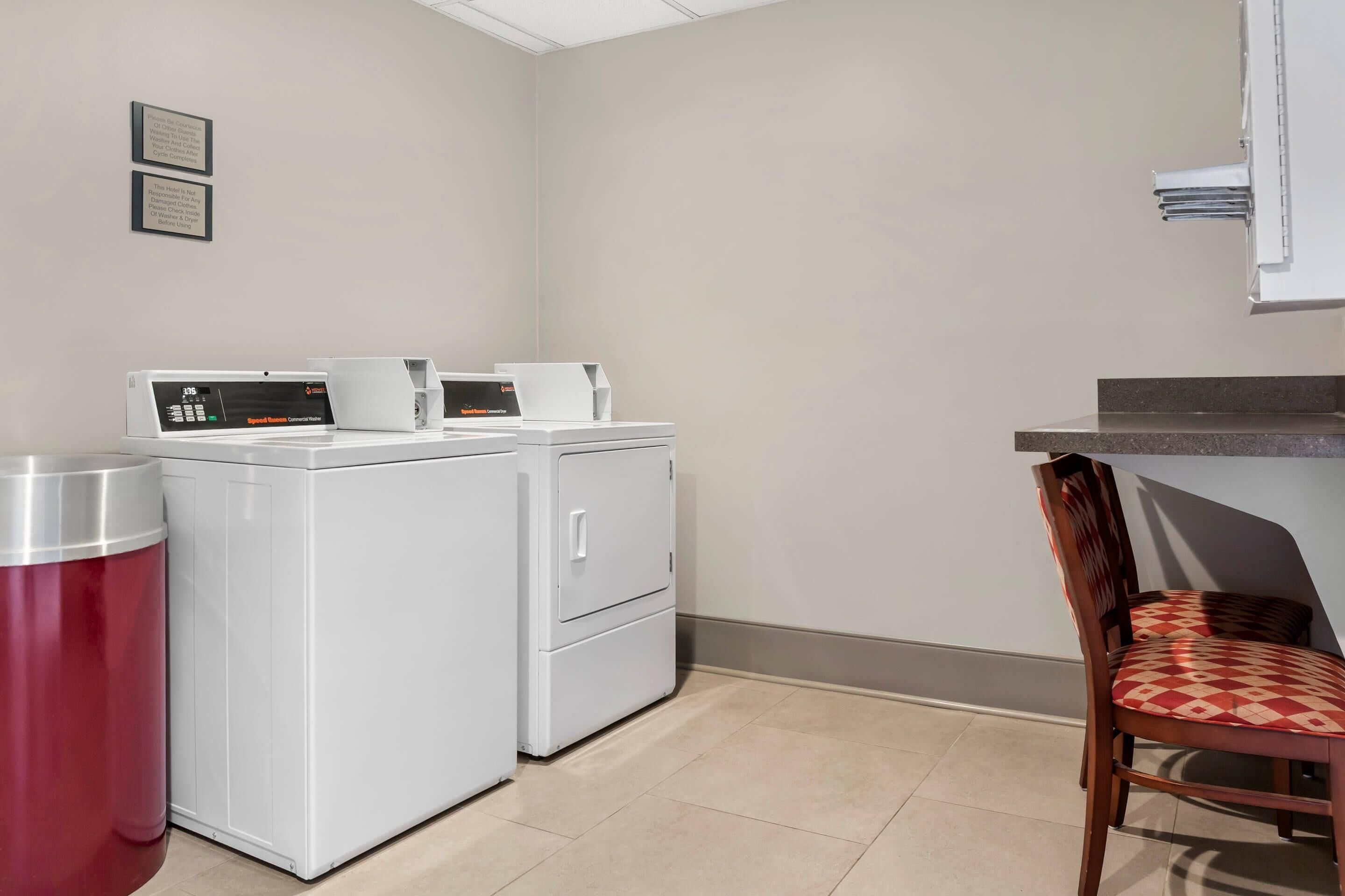A laundry area with two white washing machines, a red trash bin, and a wooden chair with a checkered cushion.