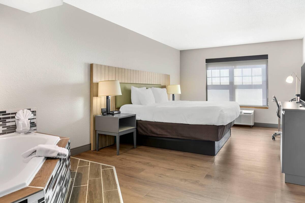 A hotel room with a bed, bedside tables, a desk, a chair, a window, and a bathroom with a bathtub.