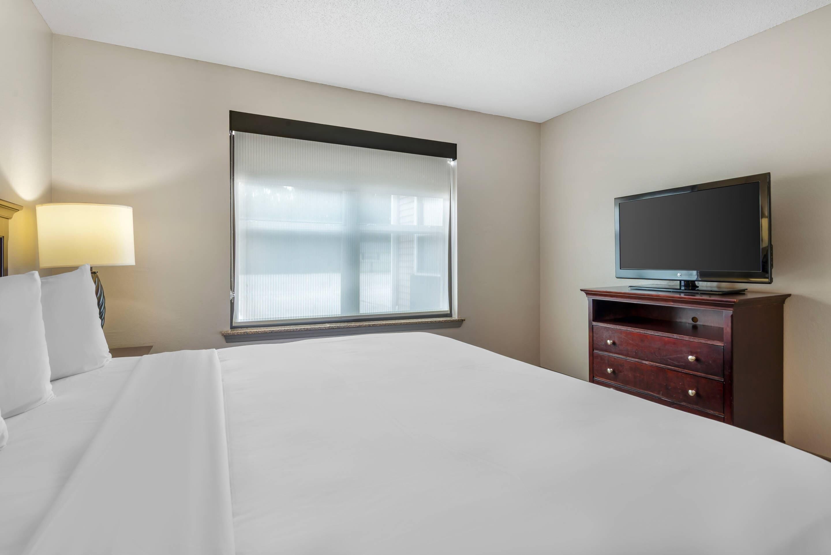 A hotel room with a bed, a television on a wooden dresser, and a window with sheer curtains.