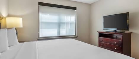 A hotel room with a bed, a television on a wooden dresser, and a window with sheer curtains.