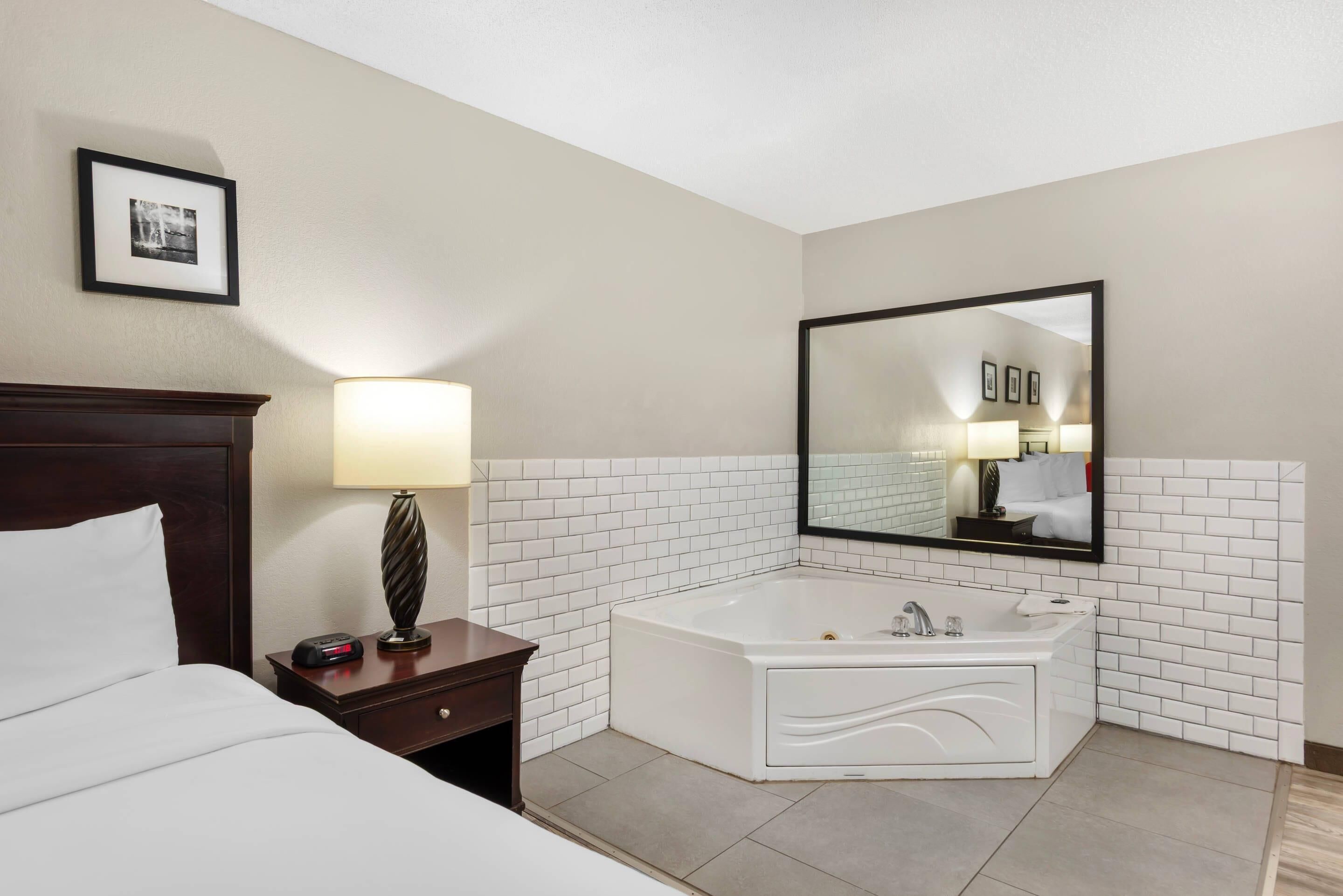 Habitación de hotel con cama, mesita de noche con lámpara, un espejo grande y un baño con azulejos blancos, una bañera y lavamanos.