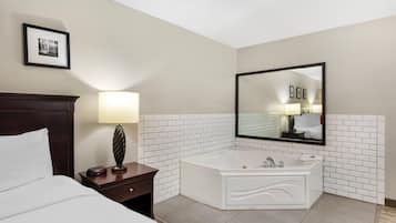 Habitación de hotel con cama, mesita de noche con lámpara, un espejo grande y un baño con azulejos blancos, una bañera y lavamanos.