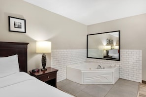 Habitación de hotel con cama, mesita de noche con lámpara, un espejo grande y un baño con azulejos blancos, una bañera y lavamanos.