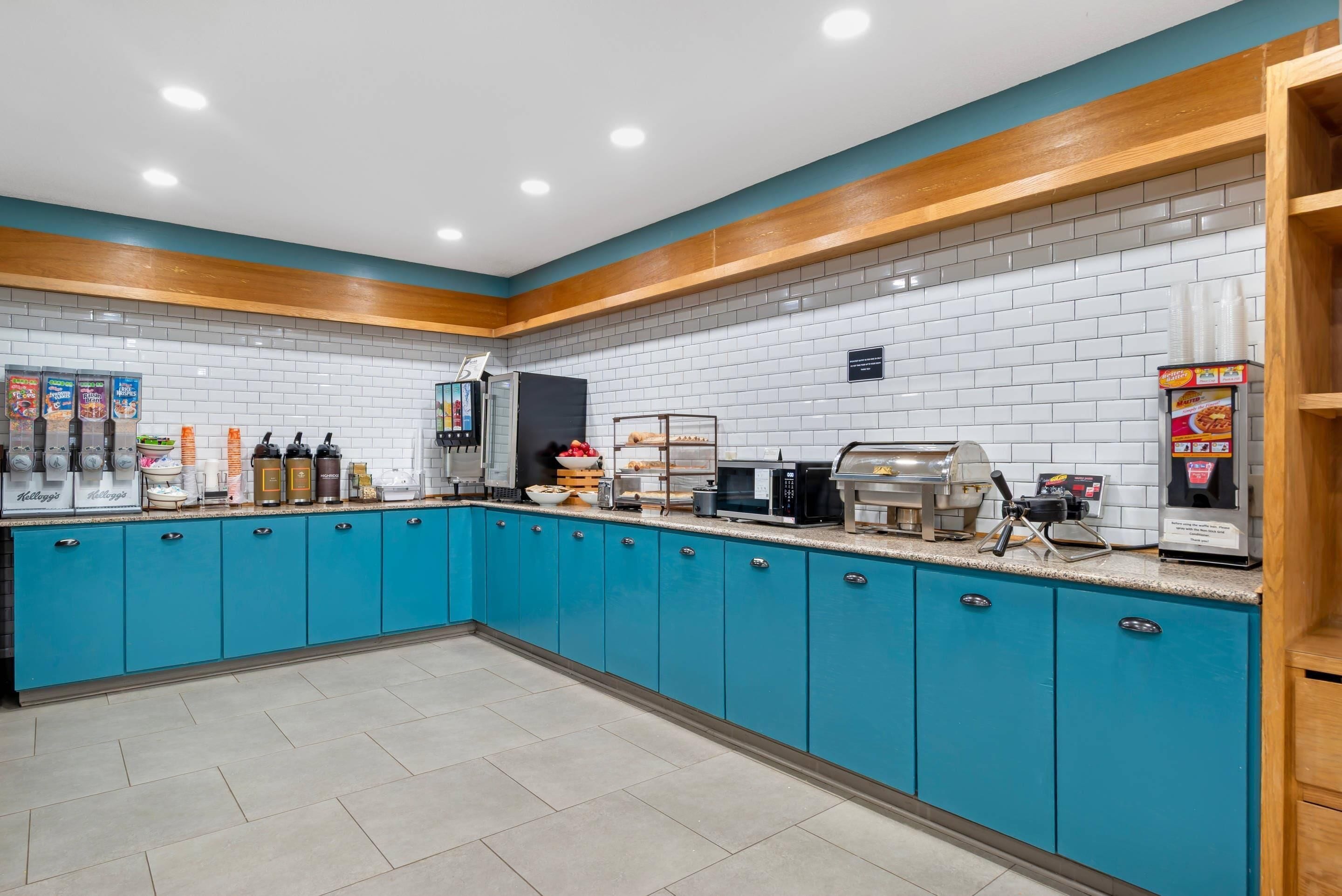 A kitchen area with teal cabinets, white tiled backsplash, and various appliances including a coffee machine, microwave, and toaster.
