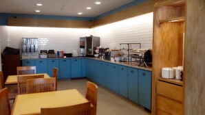 A small, modern kitchen with blue cabinets, a coffee machine, and a microwave. There are wooden chairs and a table, and a wooden cabinet with white containers on top. - Country Inn & Suites by Radisson, Hiram, GA (Hiram)