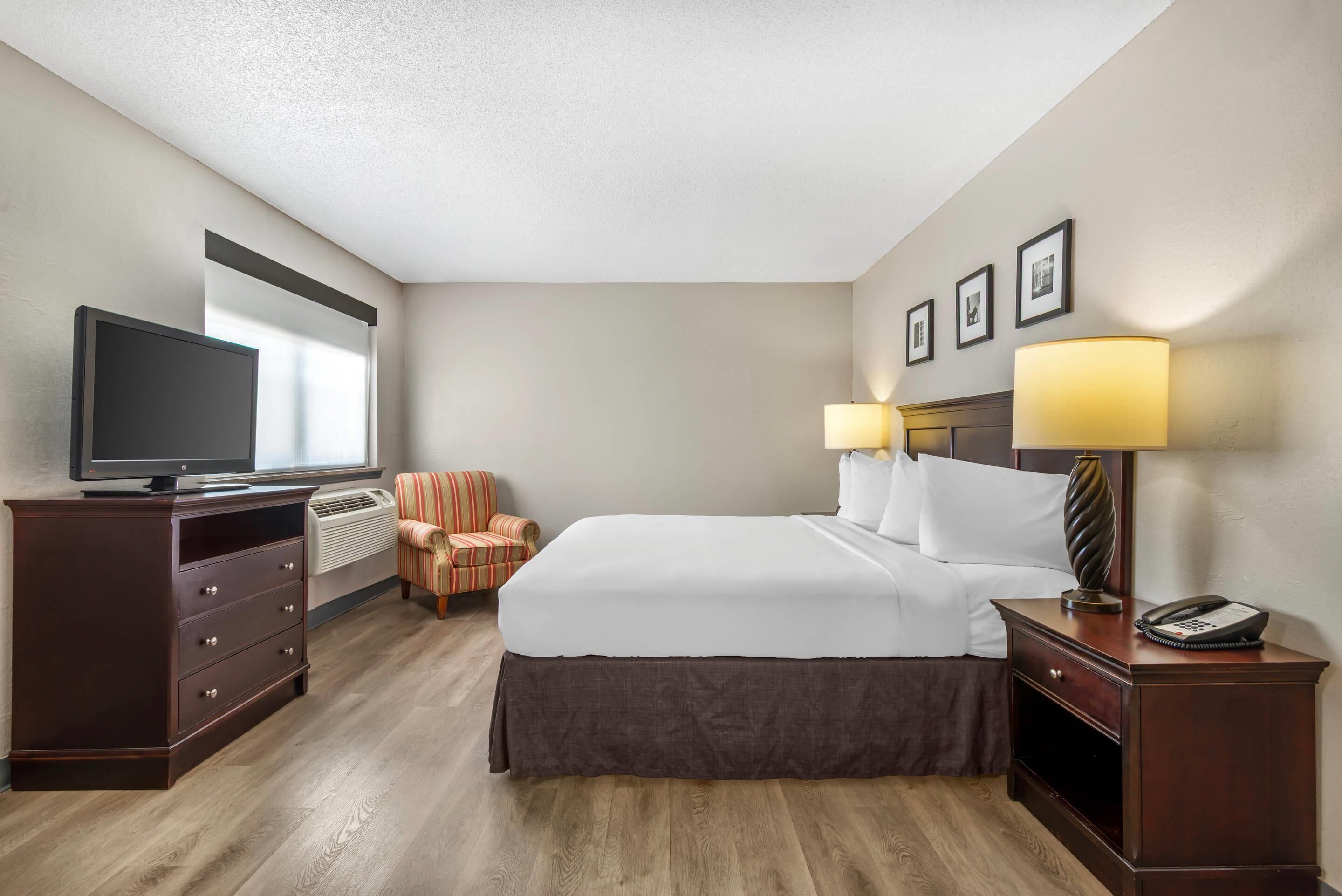A hotel room with a bed, a television, a chair, a desk with a lamp, and a phone.