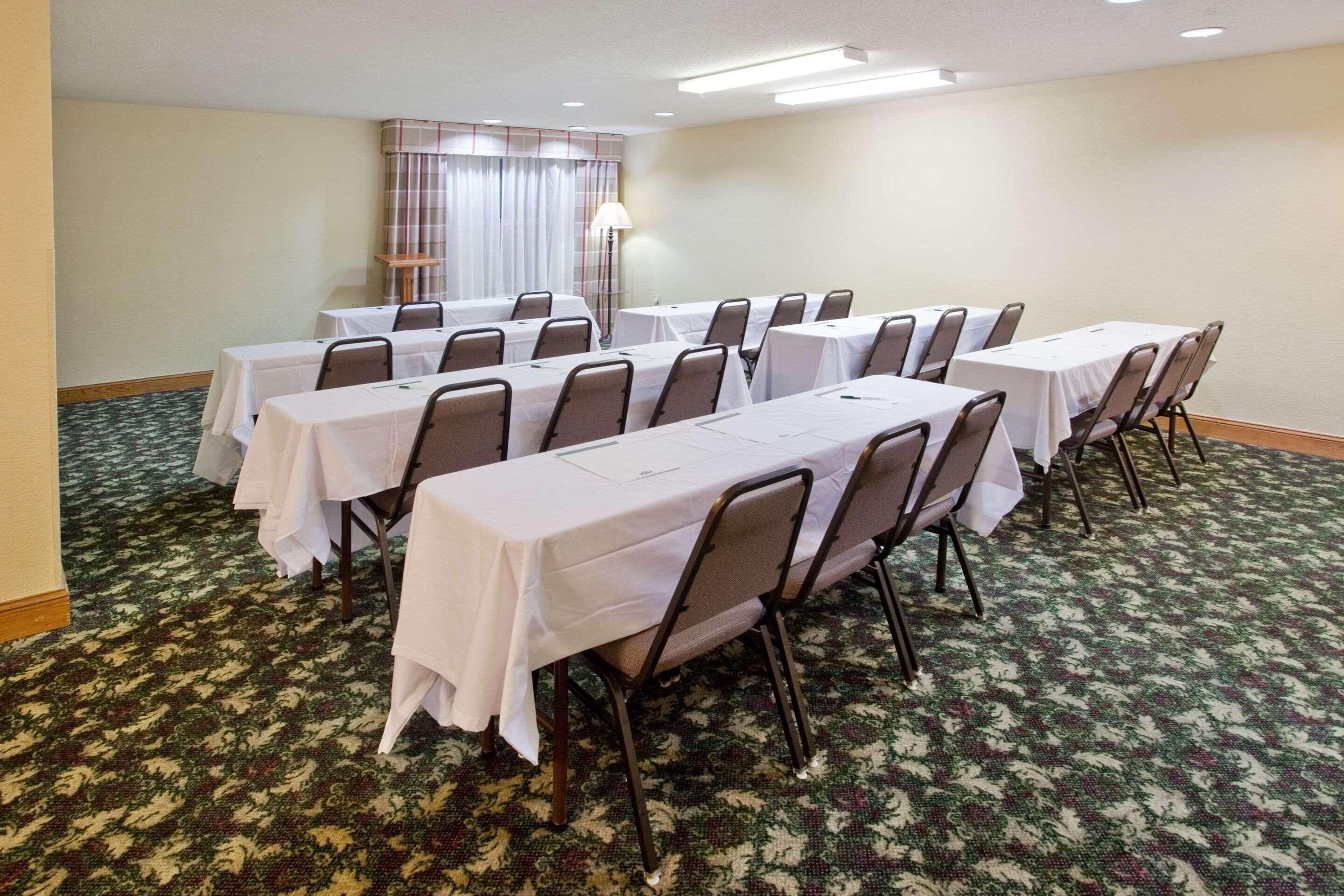 Un salón de conferencias con varias mesas y sillas, piso alfombrado y un ventanal con cortinas.