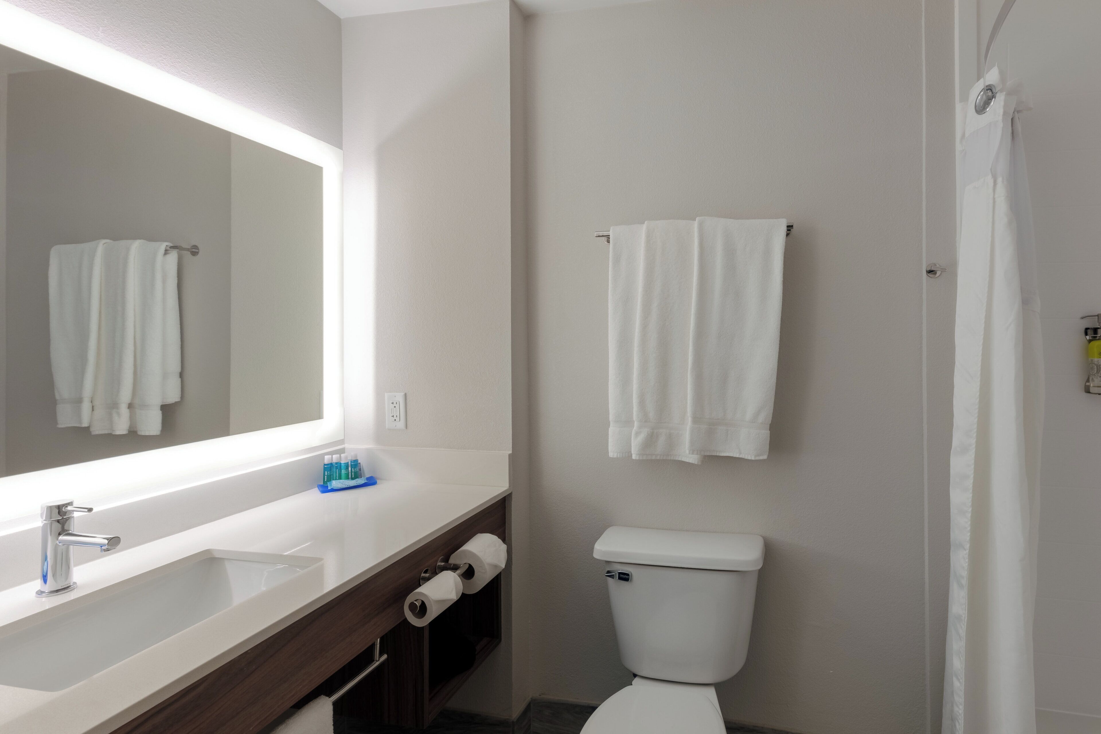 A bathroom with a white sink, mirror, and toilet. There are two rolls of toilet paper and a blue bottle on the counter.