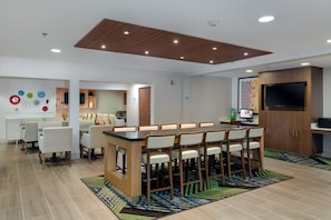 Una habitación de hotel moderna con zona de bar, asientos, un televisor de pantalla plana y letrero de centro de negocios.