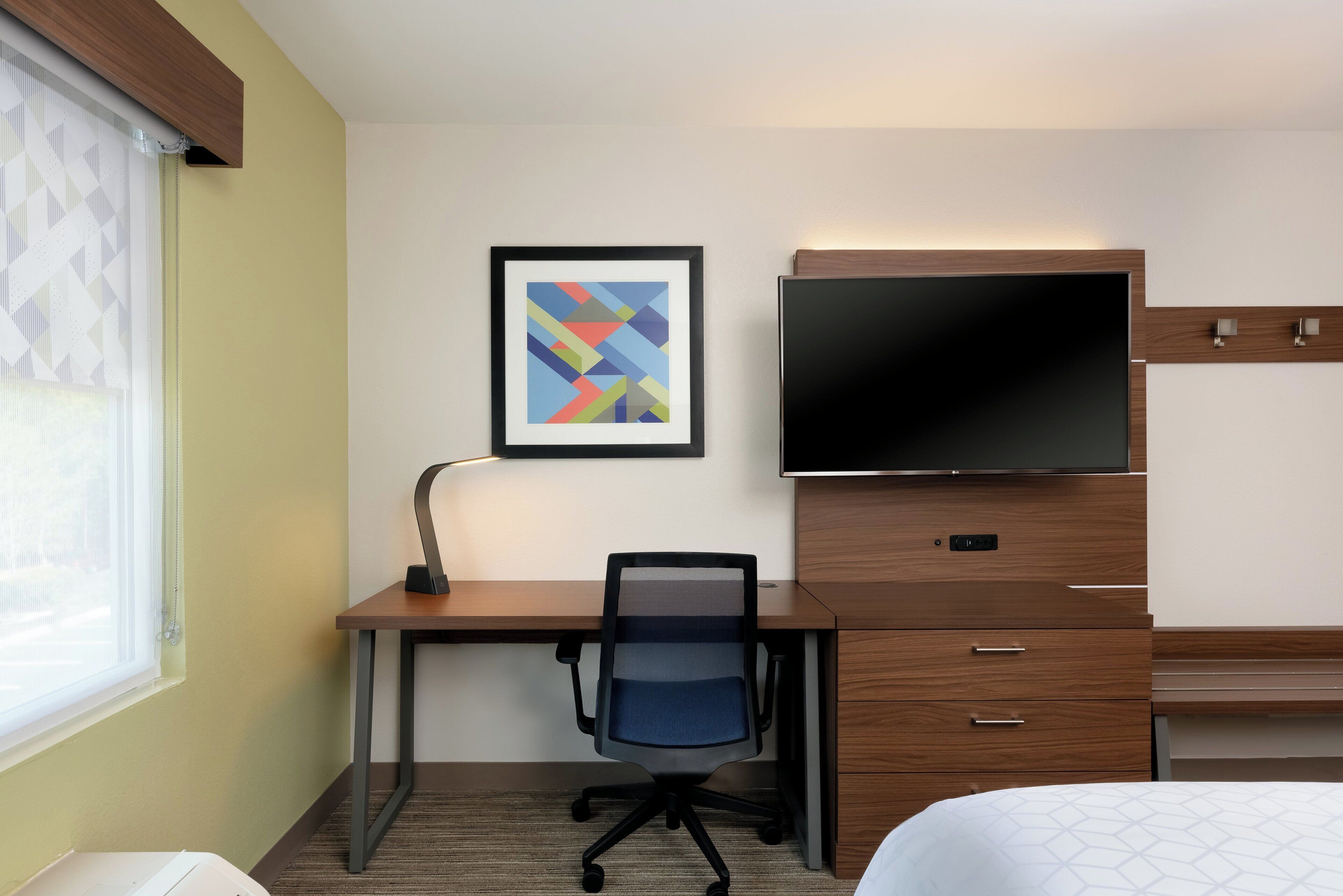 A hotel room with a desk, chair, television, and a framed artwork on the wall.