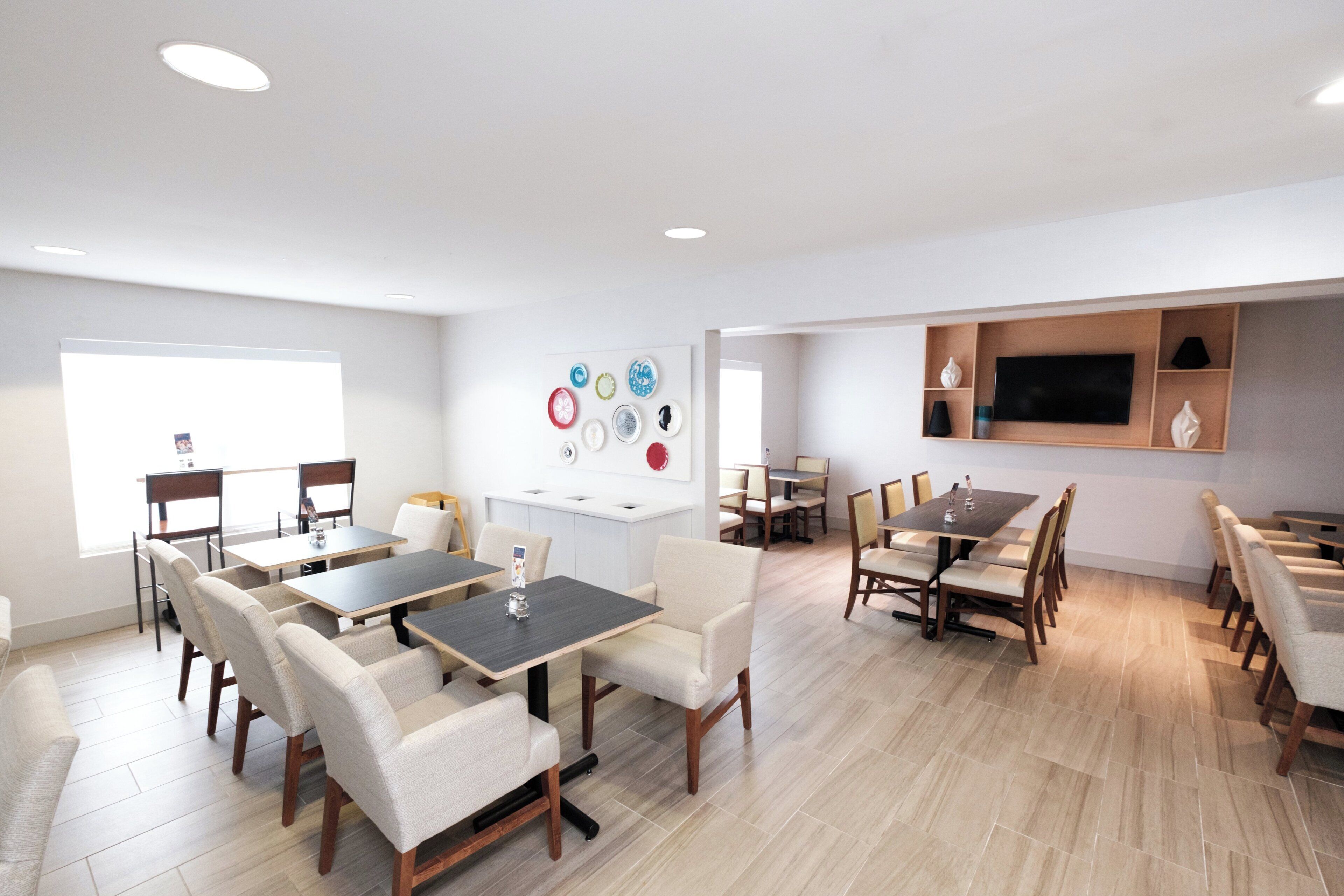 A modern conference room with multiple tables and chairs, a flat-screen TV mounted on the wall, and a decorative wall art piece.