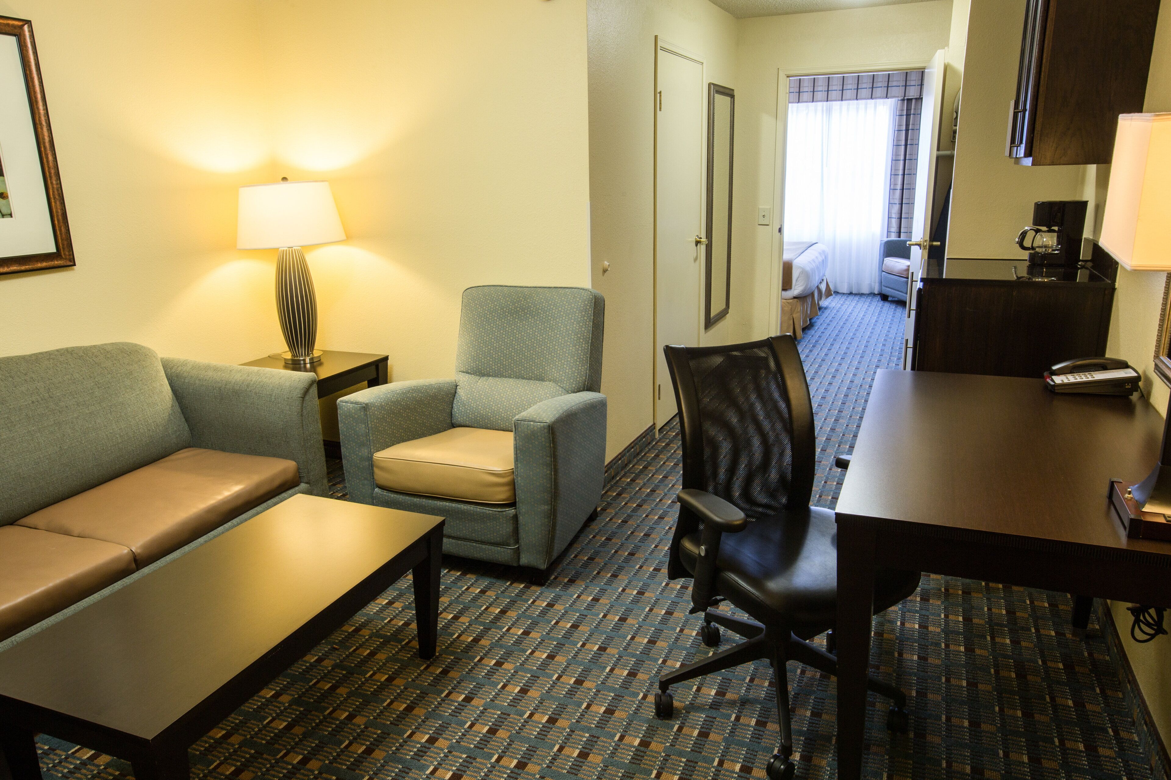 A hotel room with a sofa, armchair, desk, and a small table. There is a lamp on the armchair and a coffee machine near the desk.