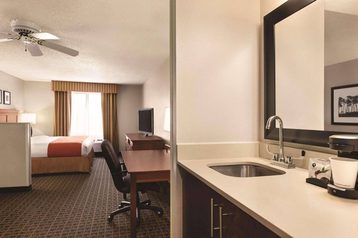 Habitación de hotel con cama, escritorio, silla y una kitchenette con lavabo y cafetera.
