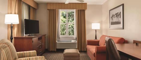 Habitación de hotel con televisión, sofá, sillón, mesa, ventana con cortinas, ventilador de techo y un cuadro en la pared.