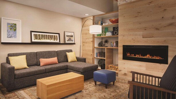 Een moderne woonkamer met een modulaire bank, een houten salontafel, een blauwe poef, een open haard en een boekenkast met diverse objecten.