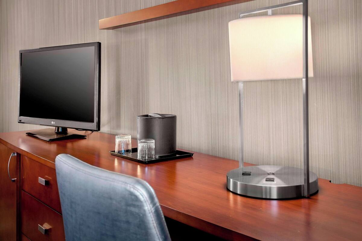 A hotel room with a wooden desk, a flat-screen TV, a lamp, and a trash can.