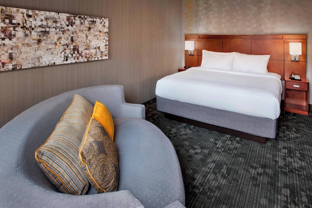 A modern hotel room with a large bed, a curved gray sofa with patterned cushions, a decorative wall art piece, and a patterned carpet.