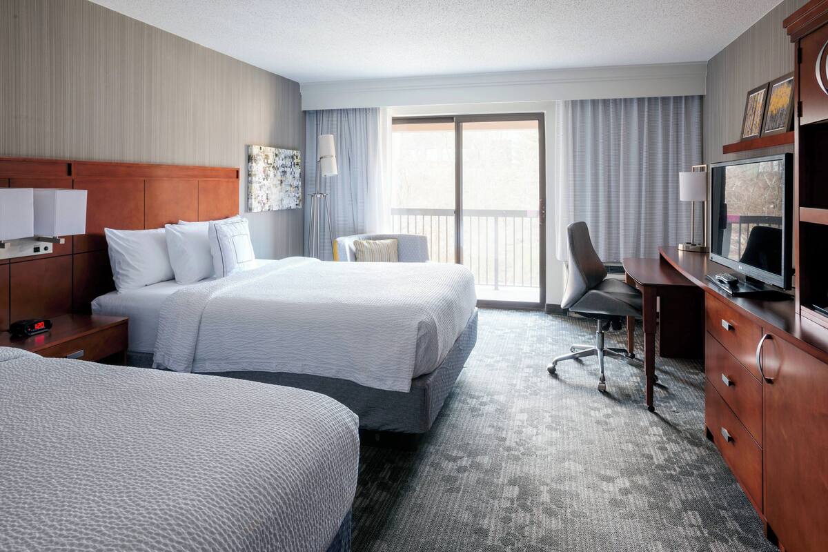 A hotel room with two beds, a desk with a computer, a chair, and a large window with curtains.