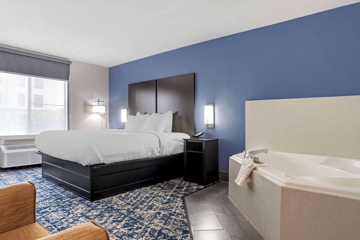 A hotel room with a bed, bedside table, lamp, and a large bathtub.