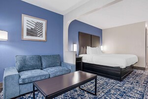 A hotel room with a blue wall, a bed with white bedding, a blue sofa, a dark coffee table, and a framed picture on the wall.