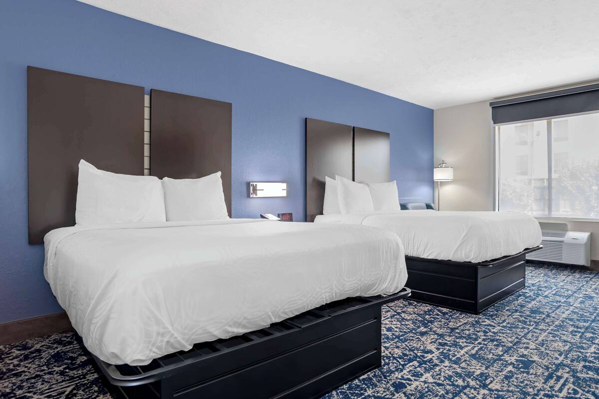 A hotel room with two beds, a large window, a wall clock, and a nightstand with a lamp.
