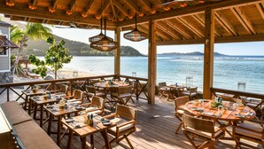 Una terraza de madera con mesas y sillas, con vista a la playa y al océano.