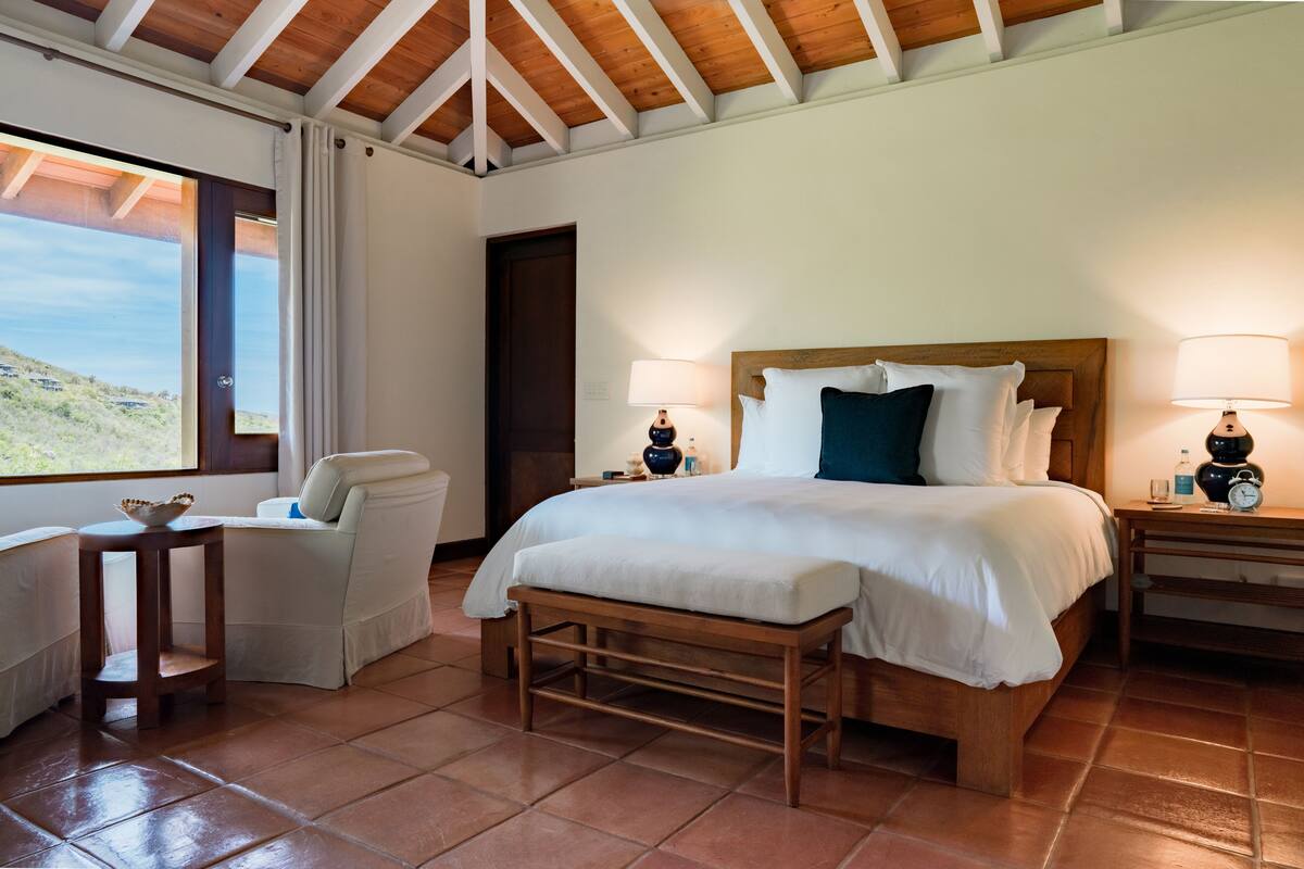 Une chambre à coucher avec un grand lit, deux tables de chevet, un banc, une chaise et une fenêtre donnant sur le paysage.