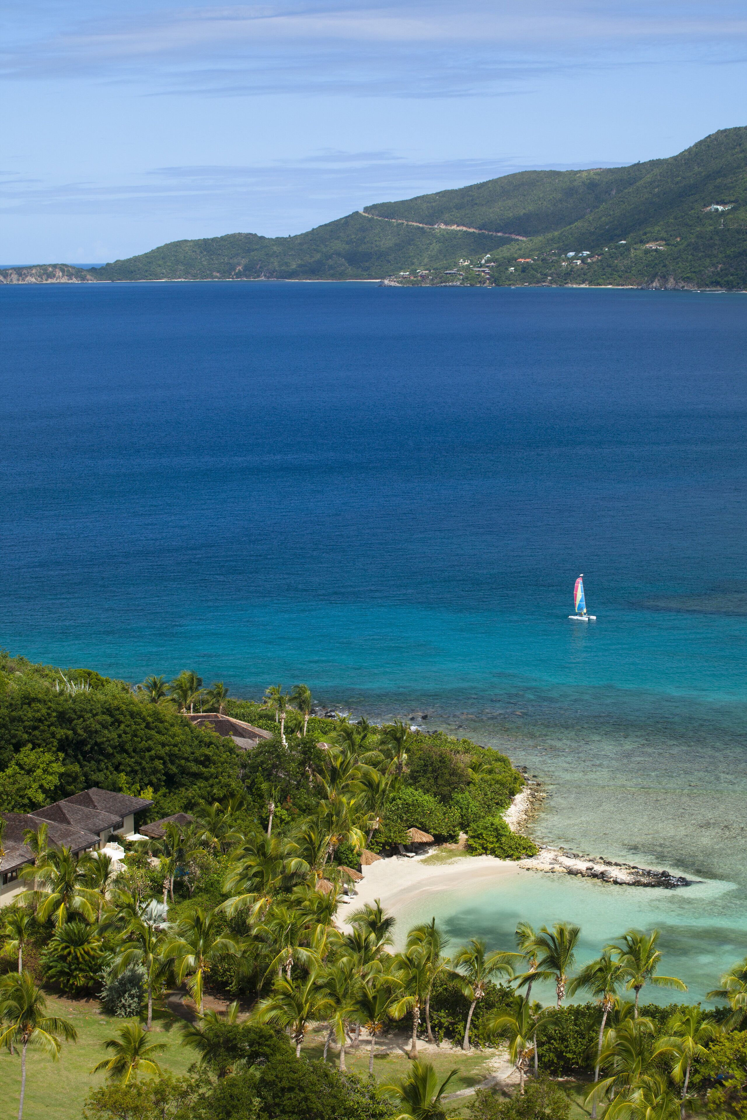 Vista costera con un velero en el agua, una playa y abundante vegetación.