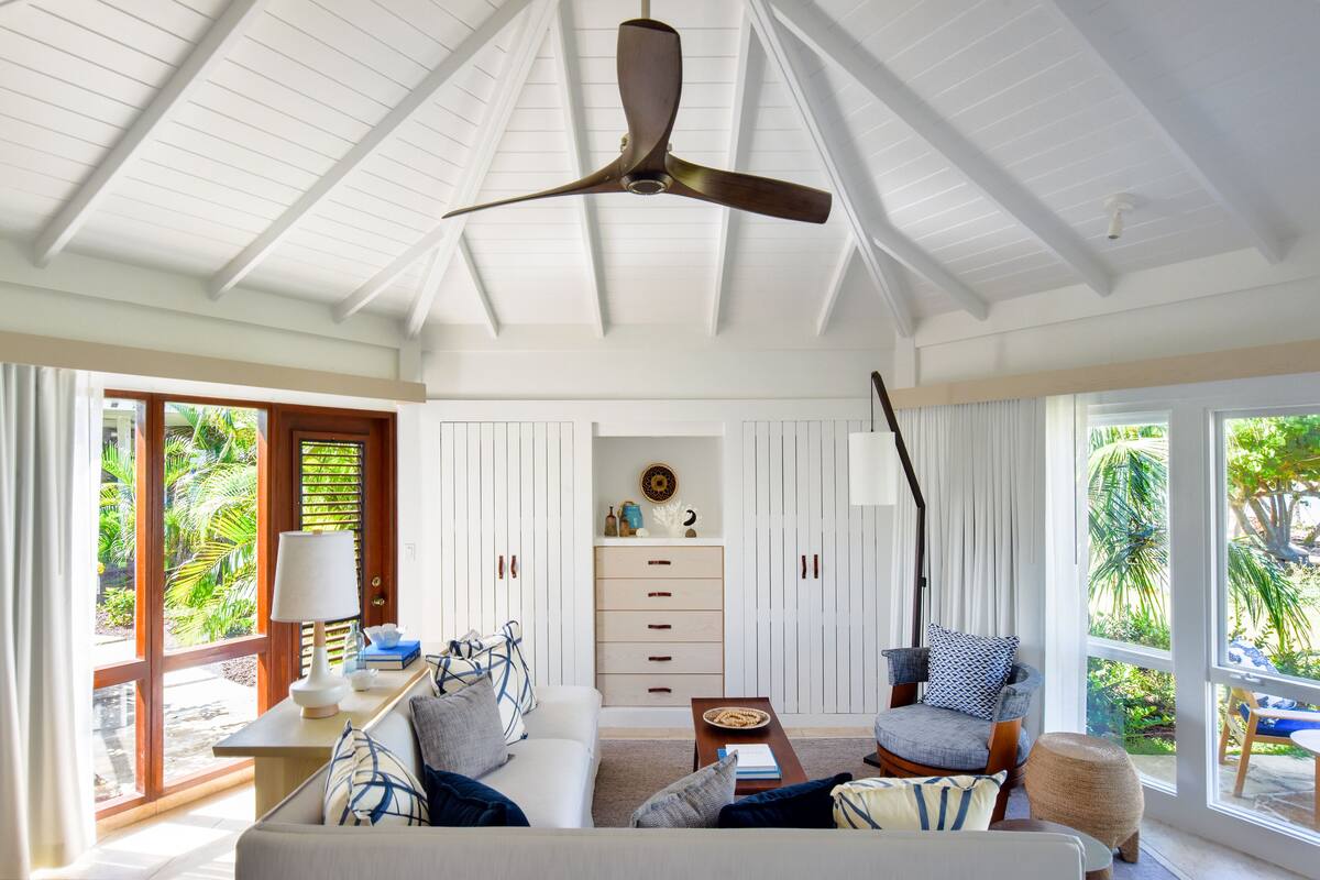 Un salon spacieux doté d’un ventilateur de plafond, d’un canapé, d’une table basse et d’un fauteuil. Une fenêtre offre une vue sur la verdure environnante.
