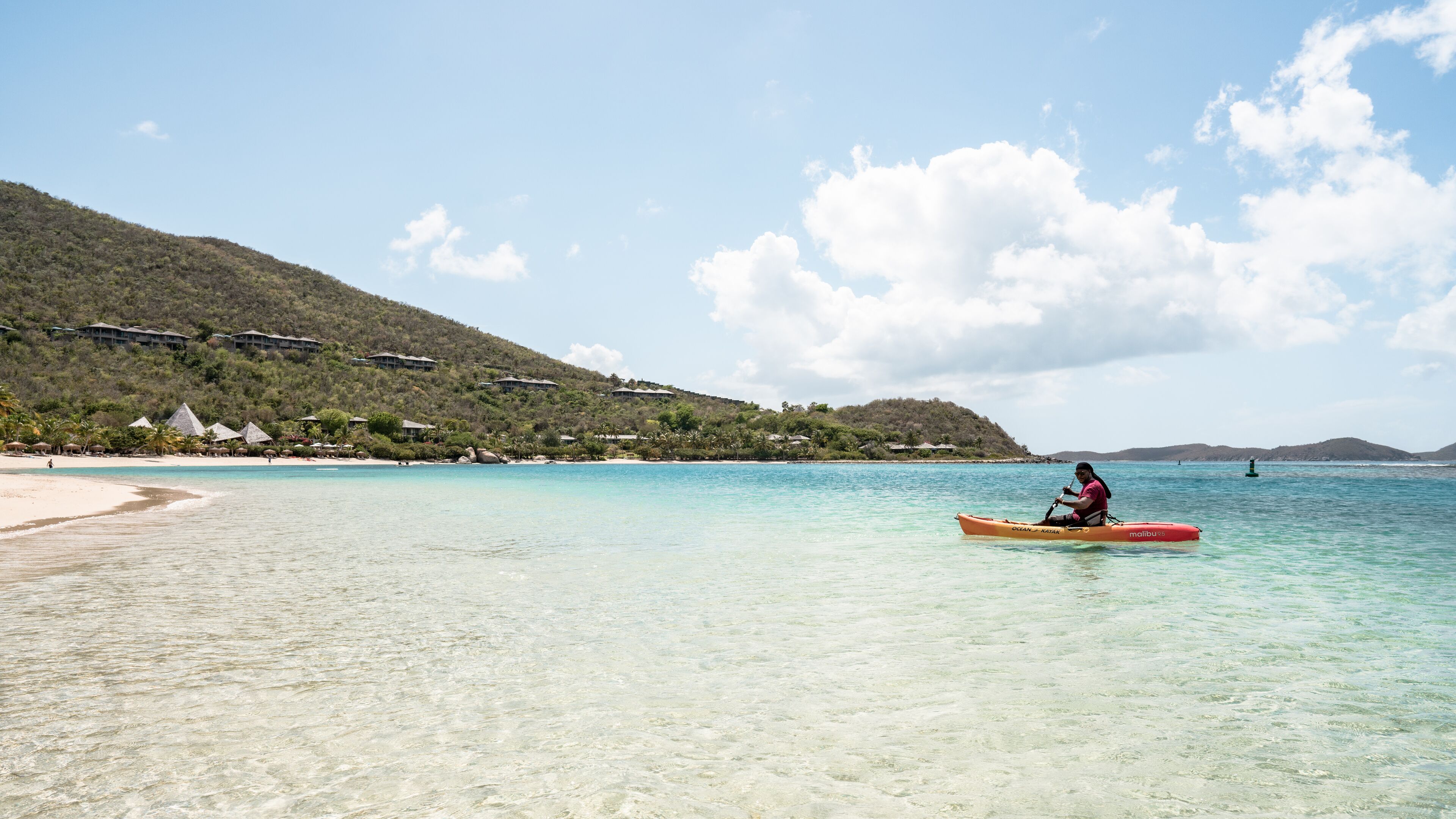 A clear beach with a person kayaking, a sandy shore, and a backdrop of hills with buildings.