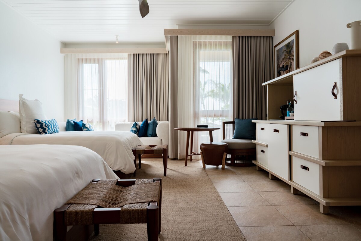 A hotel room with two beds, a desk, a chair, a small table, and a cabinet.