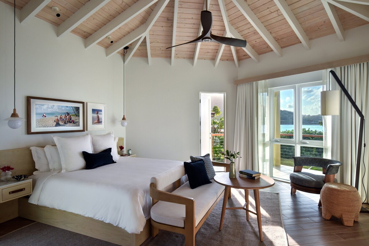 A spacious bedroom with a large bed, a ceiling fan, a view of the outdoors, and a dining area with a table and chairs.