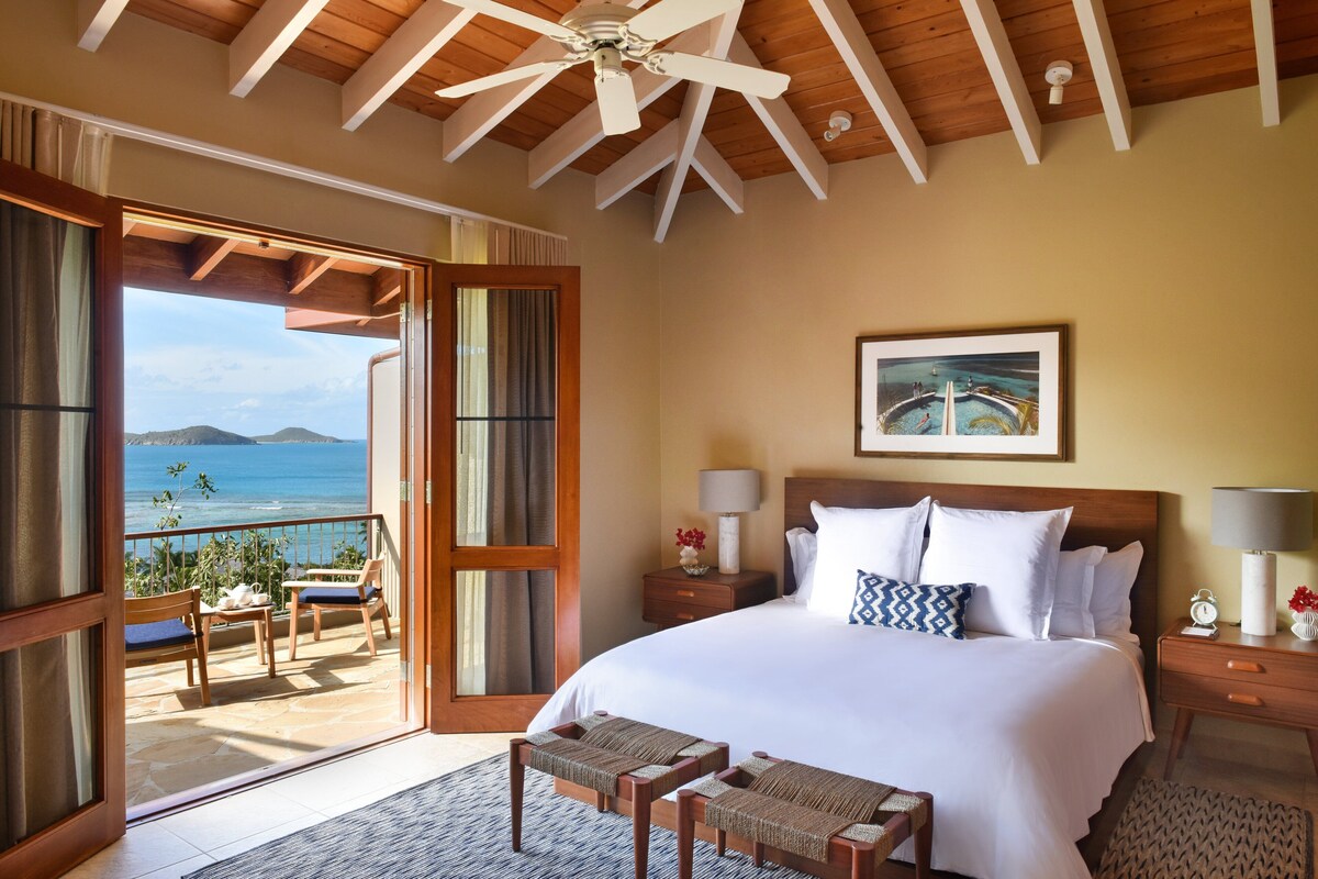 A bedroom with a large bed, a balcony with a view of the ocean, and a ceiling fan.