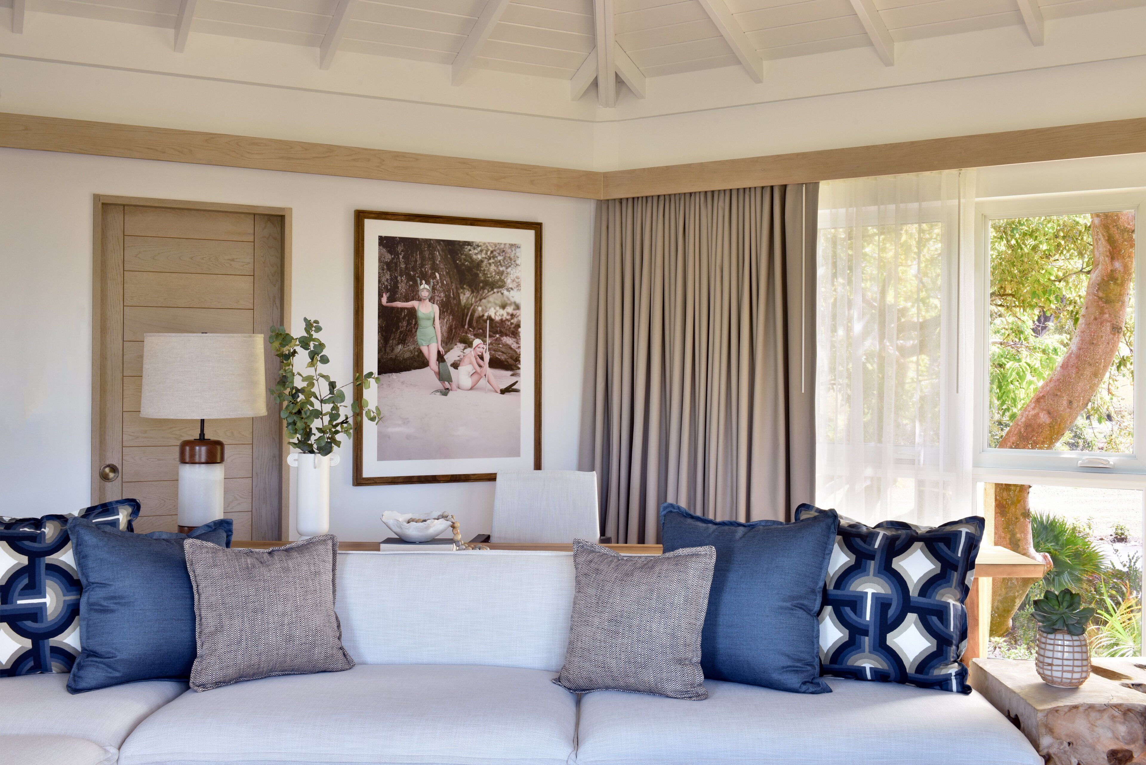 Un salón moderno con un sofá blanco, almohadones estampados en azul, una fotografía enmarcada, una lámpara y una ventana con cortinas.