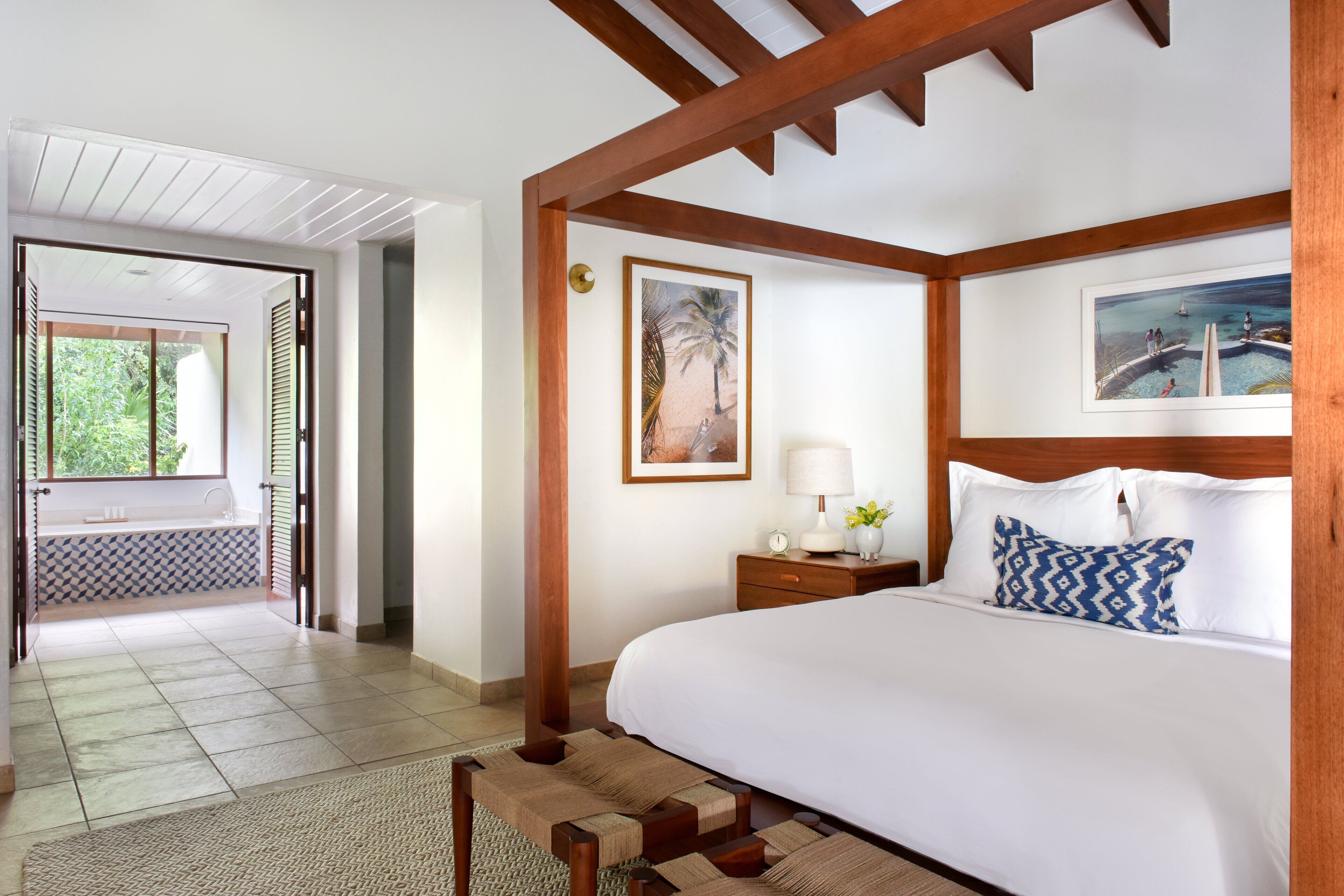 A hotel room with a large bed, a nightstand with a lamp, a framed picture on the wall, and a view of the outdoors through an open door.