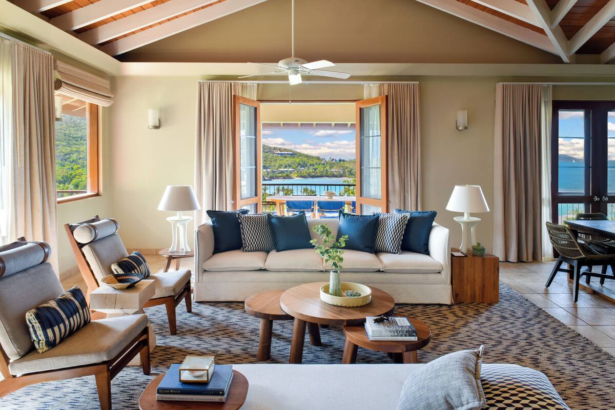 Un salon spacieux comprenant un canapé, deux fauteuils, une table basse et un coin repas avec une table et des chaises. Une grande fenêtre offre une vue sur un paysage verdoyant.